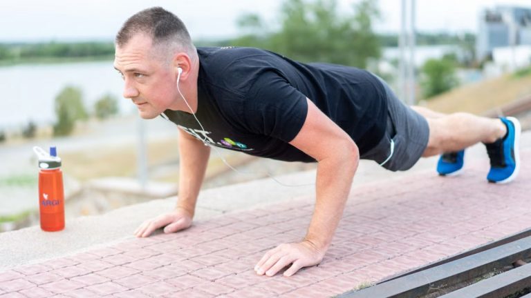 Adult male exercising outdoors performing push-ups with earphones and water bottle.